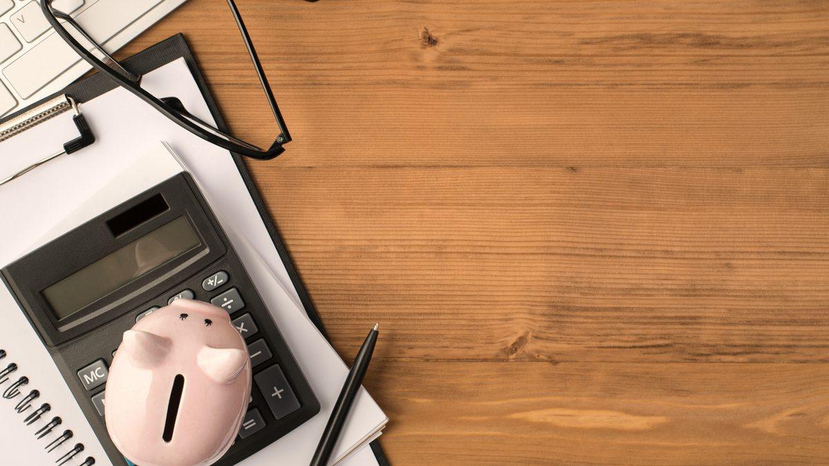 Une tirelire rose posée sur une calculatrice et un porte-documents, à côté d'une paire de lunettes, sur un bureau en bois