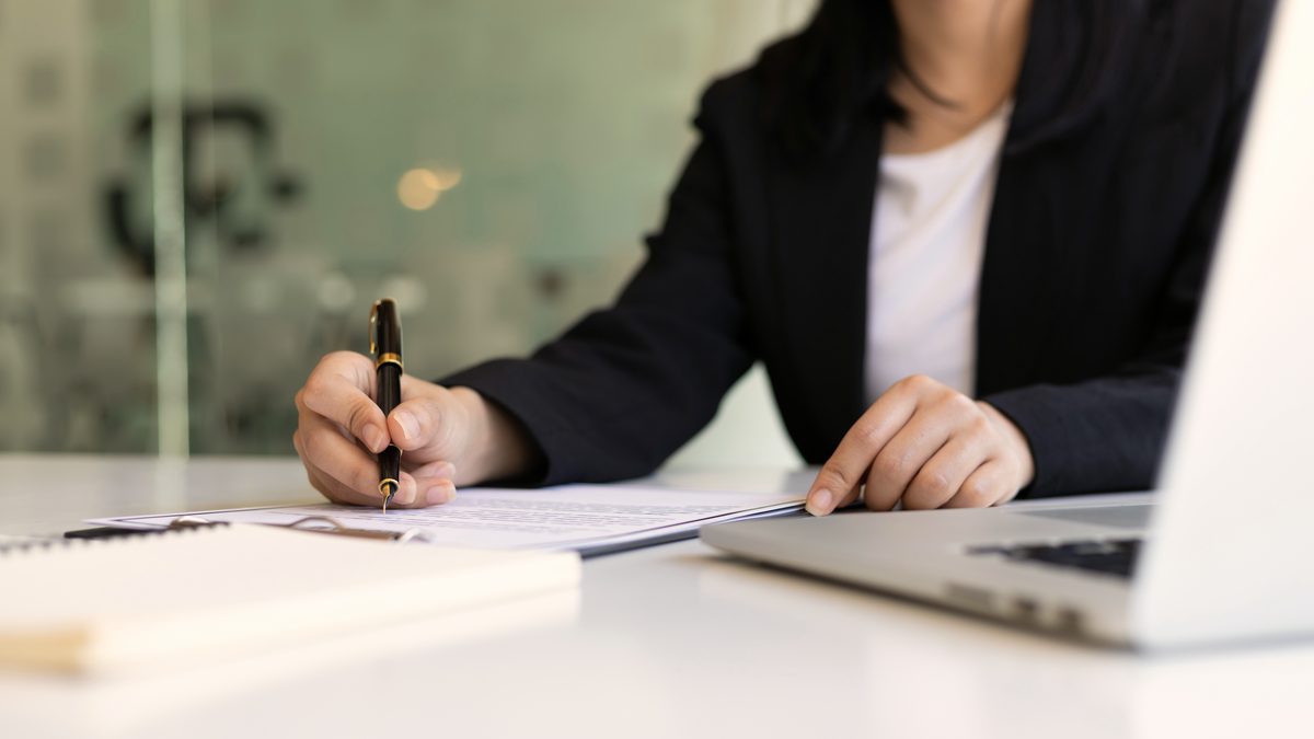 Femme en veste sombre signant un document au stylo sur un bureau clair, à côté d'un carnet et d'un ordinateur portable
