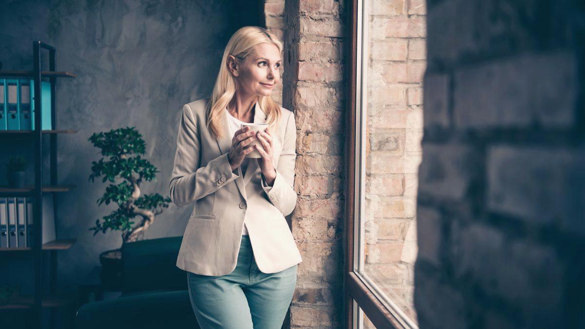 Professionnelle debout près d'une fenêtre, une tasse à la main, prenant un moment de réflexion