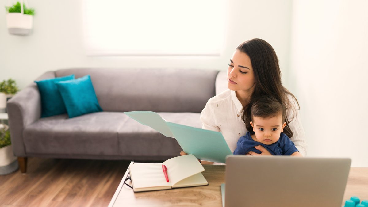 Une maman travaille sur son ordinateur portable à la maison, son bébé sur les genoux, un cahier ouvert devant elle