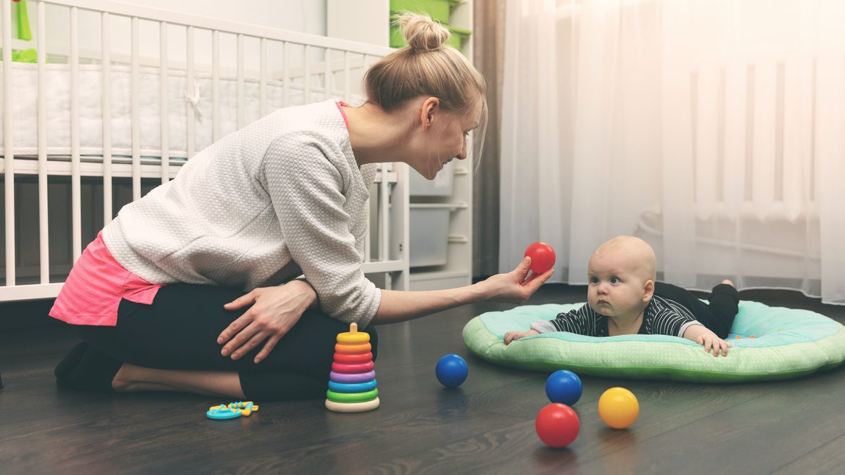 Une assistante maternelle joue avec un bébé installé sur un tapis d'éveil, au pied d'un lit à barreaux