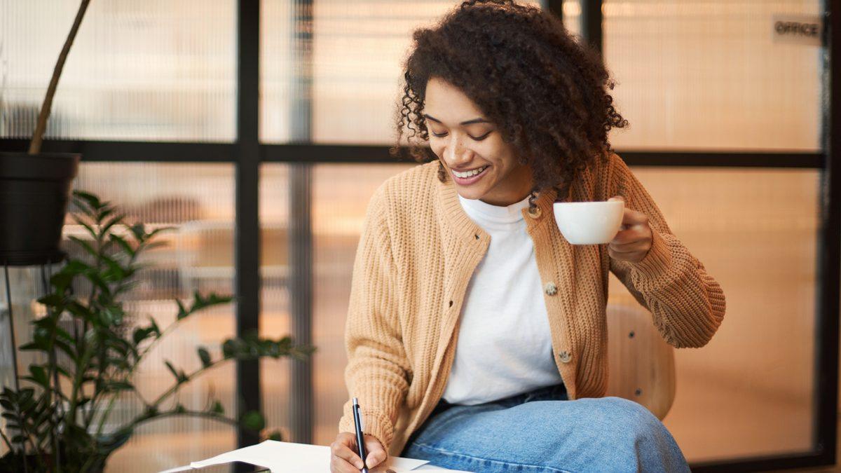 Femme souriante assise près d'une fenêtre, prenant des notes sur un document posé sur ses genoux