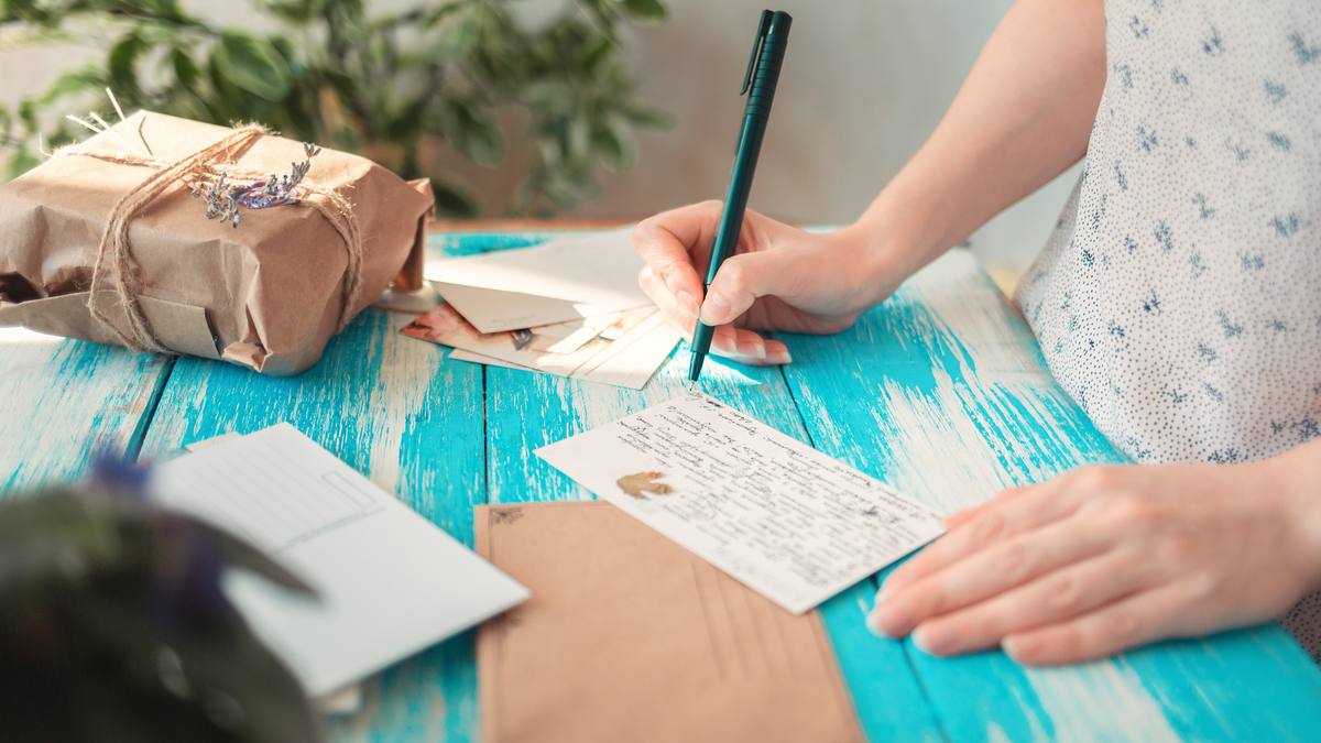 Main de femme écrivant une lettre au crayon sur une table en bois peint, à côté d'une enveloppe kraft et d'un carnet