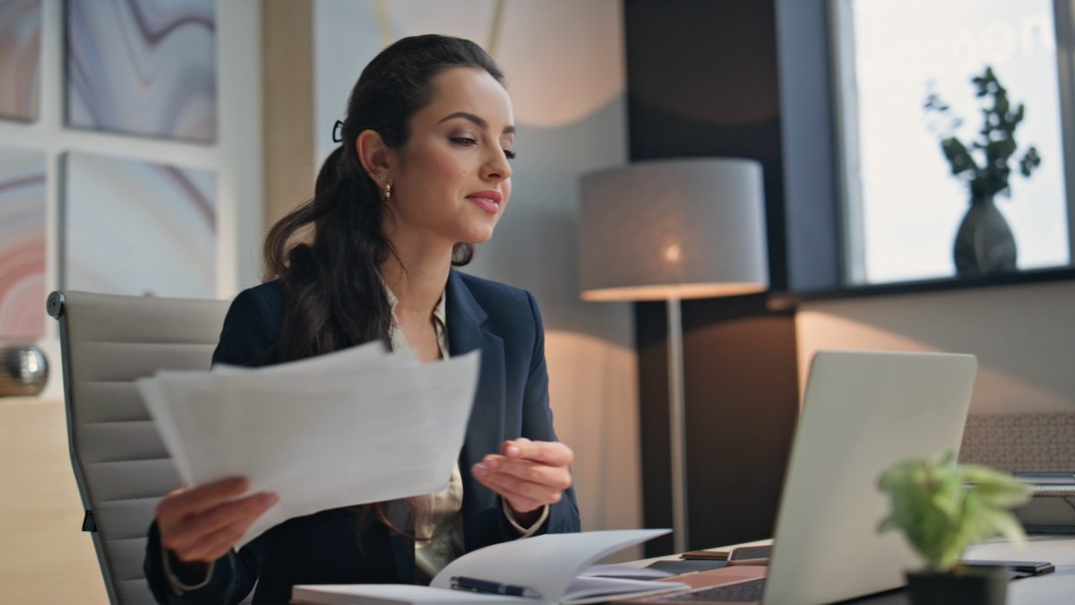 Une jeune femme en veste sombre lit une feuille devant un ordinateur portable, dans un bureau lumineux