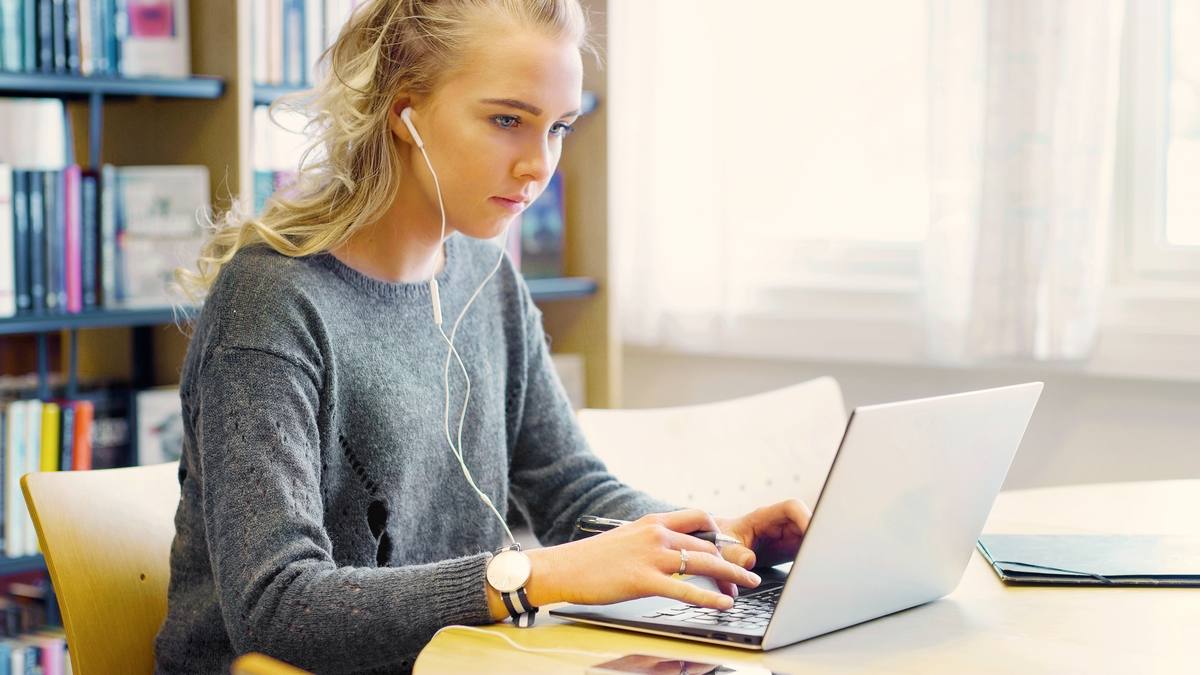 Jeune femme assise à un bureau clair, travaillant sur un ordinateur portable devant une bibliothèque