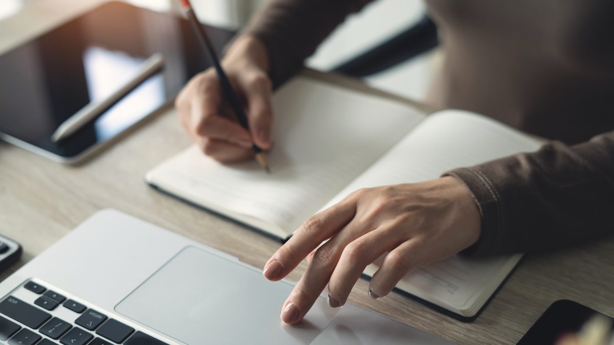 Mains d'une étudiante prenant des notes dans un carnet à côté d'un ordinateur portable ouvert, sur un bureau clair