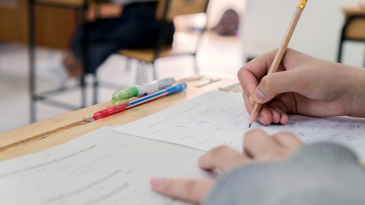 Main d'une candidate écrivant au crayon sur une copie d'examen, stylos posés sur la table