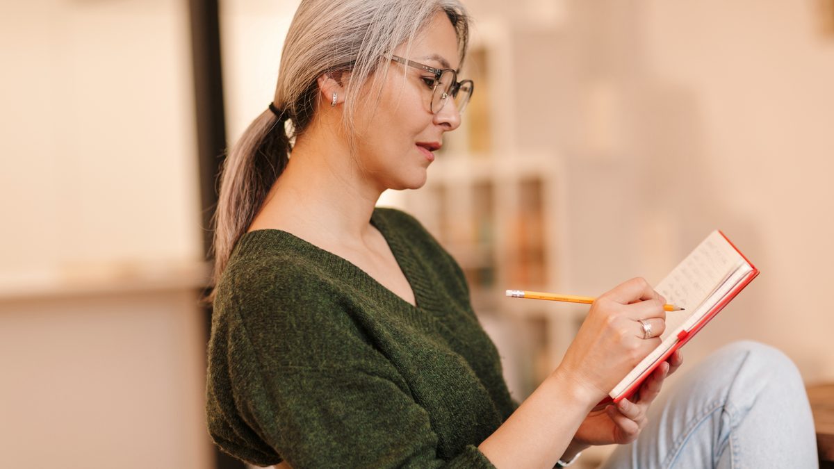 Femme à lunettes écrivant au crayon dans un carnet rouge, assise dans un salon lumineux