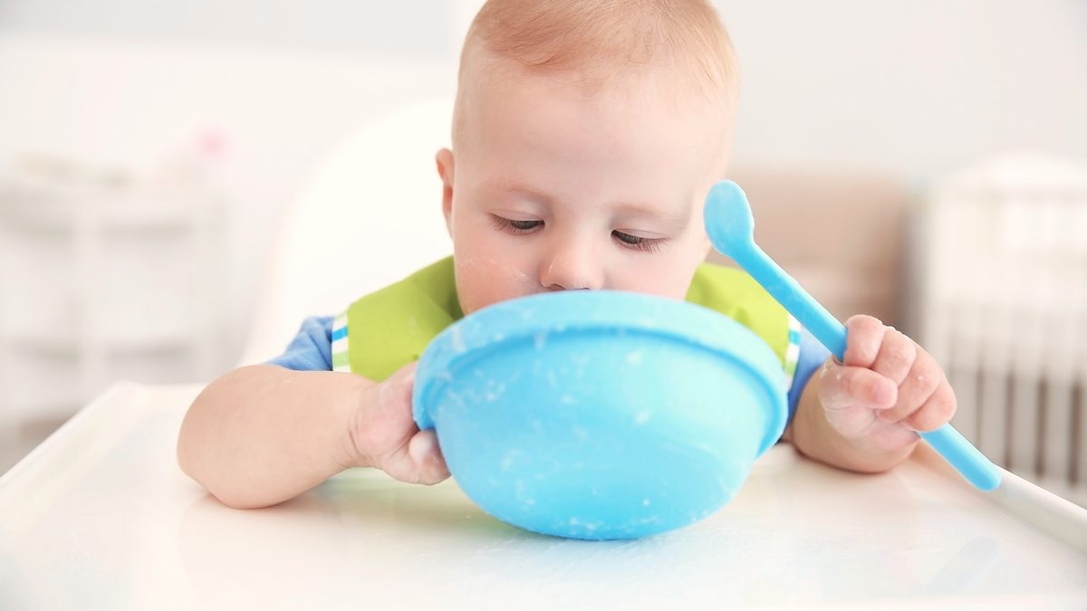 Un bébé mange à la cuillère dans un bol, installé à table