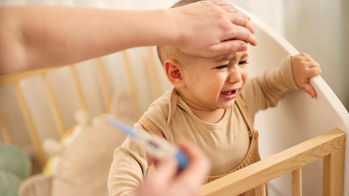 Une maman pose sa main sur le front de son bébé qui pleure dans son lit, un thermomètre à la main