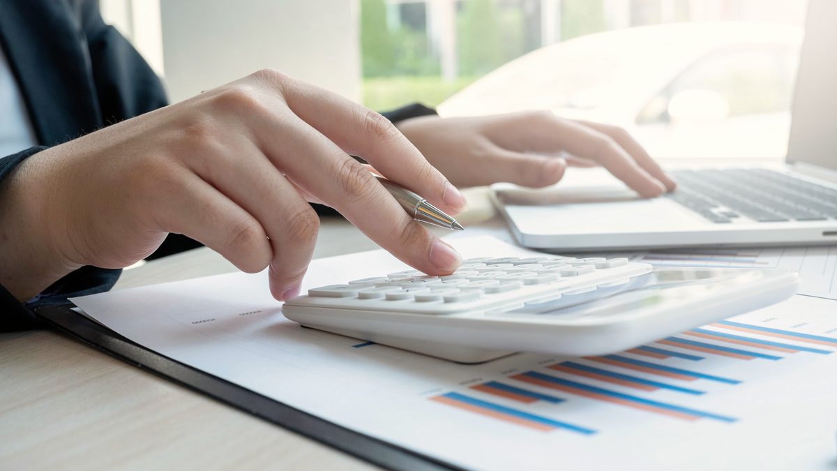 Mains d'une femme tapant sur une calculatrice posée sur des documents chiffrés, à côté d'un ordinateur portable