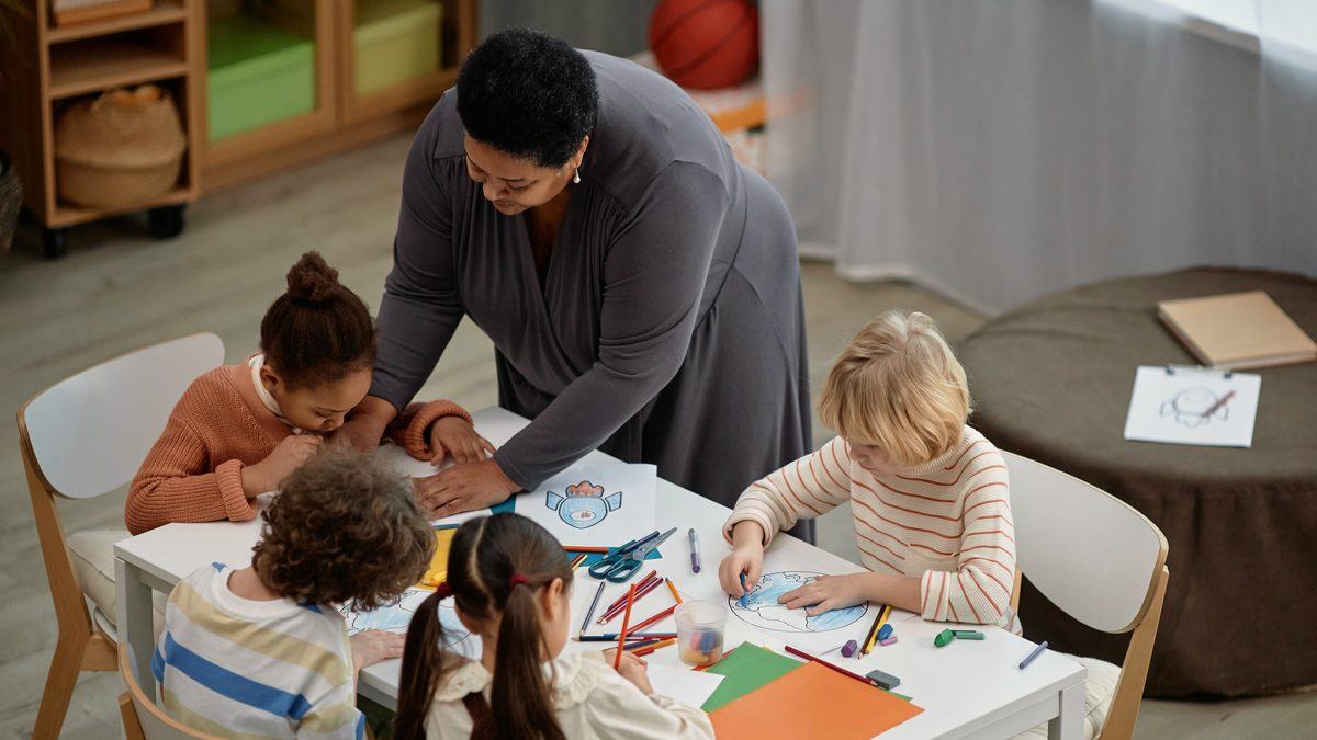 Une adulte penchée vers un groupe de jeunes enfants attablés autour d'un atelier de collage en classe maternelle
