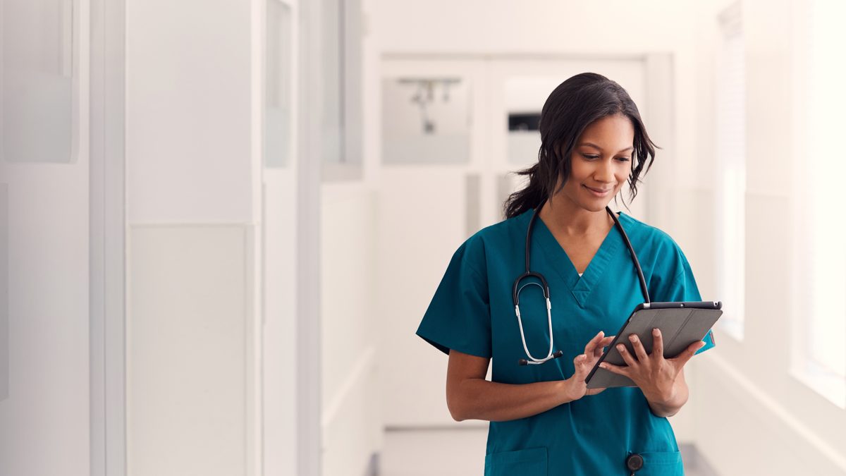 Professionnelle de santé en tenue de soins consultant une tablette dans un couloir d'établissement