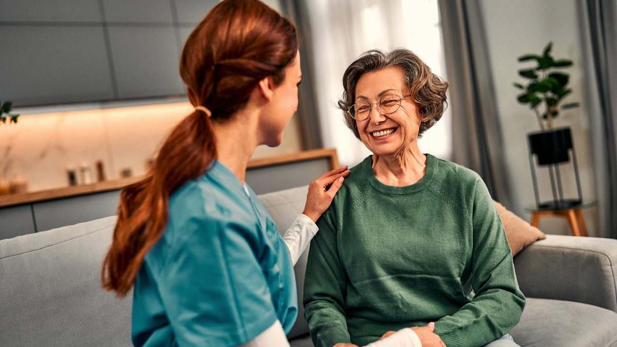 Soignante en tenue professionnelle accompagnant une personne âgée souriante dans un salon lumineux