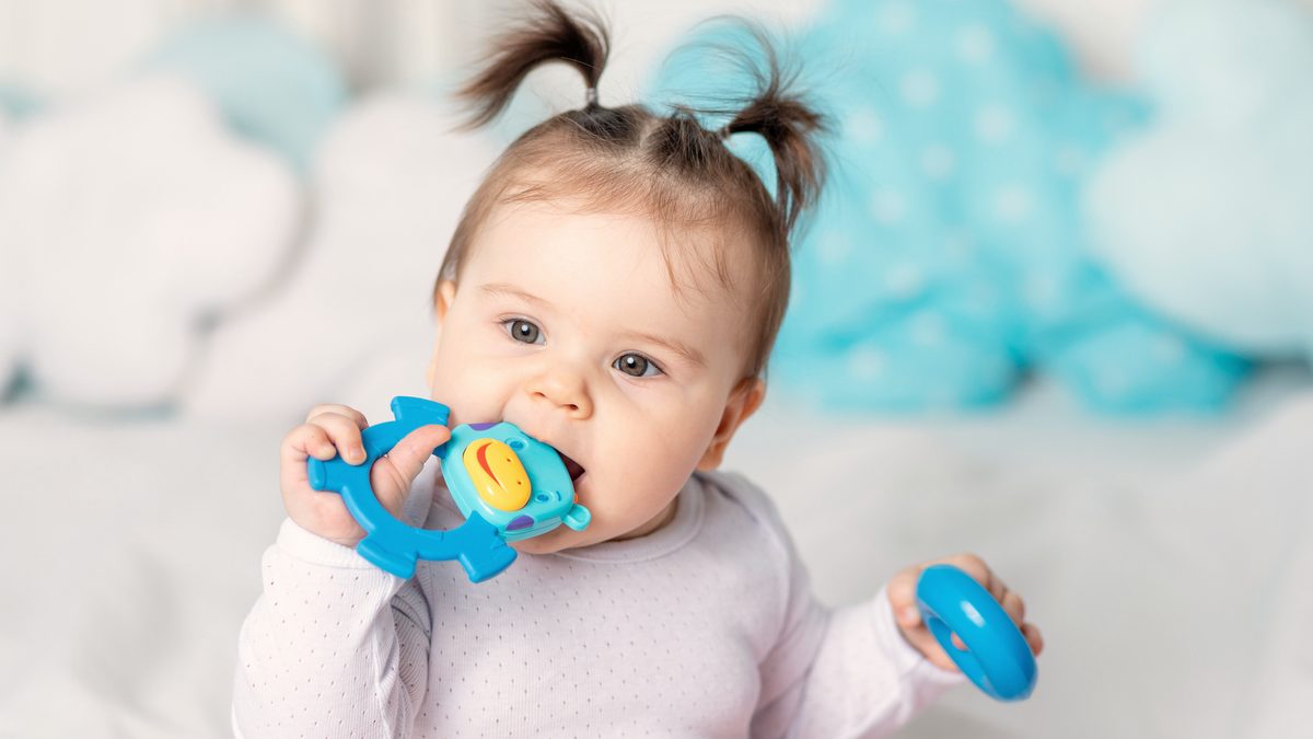 Un bébé mordille un anneau de dentition bleu, assis sur un lit