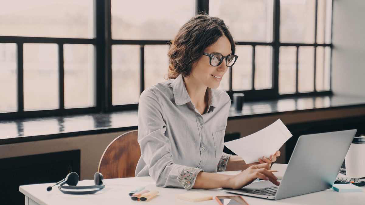 Une jeune femme à lunettes souriante consulte une feuille devant son ordinateur portable, dans un bureau lumineux