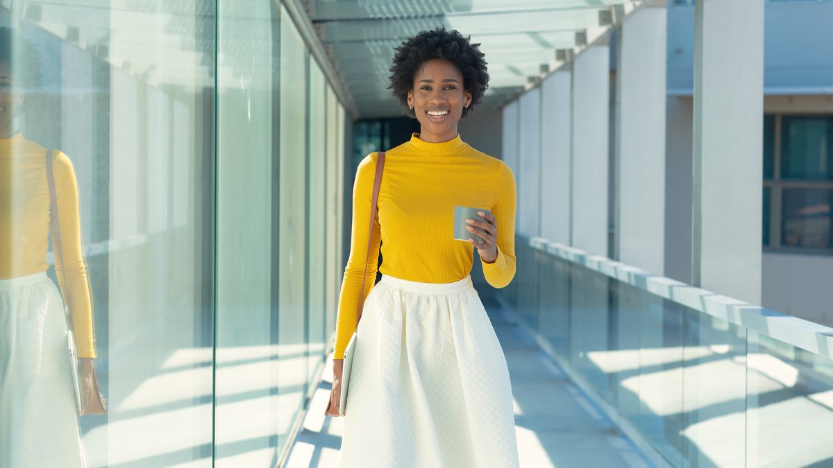 Une femme souriante marche dans un couloir lumineux, une tasse à la main