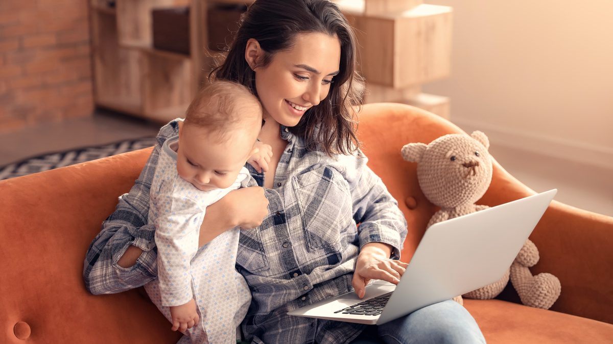 Une jeune maman installée sur un canapé orange travaille sur son ordinateur portable, son bébé dans les bras