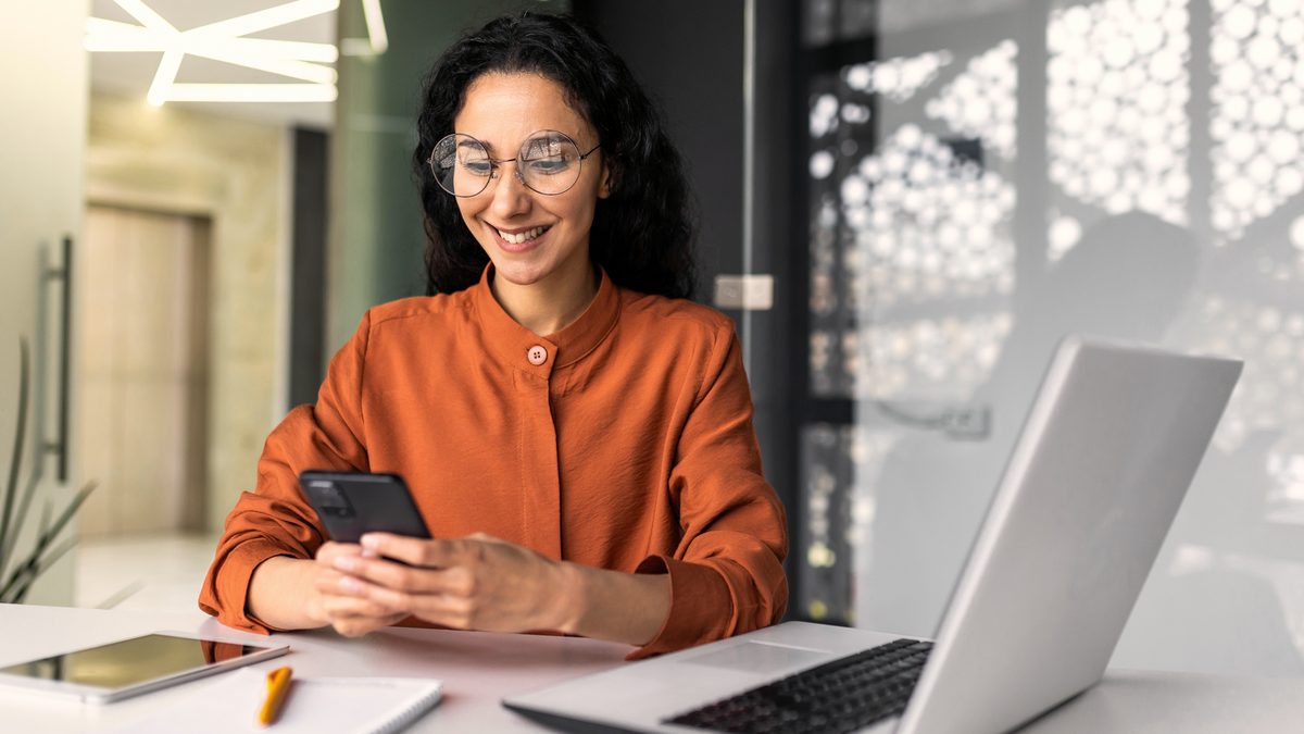 Une jeune femme souriante consulte une bonne nouvelle sur son téléphone, devant son ordinateur portable