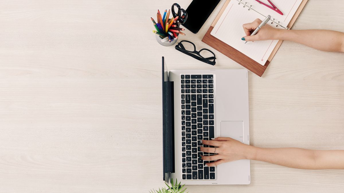 Vue de dessus d'un bureau clair : une main écrit sur un agenda à spirale, l'autre tape sur un ordinateur portable