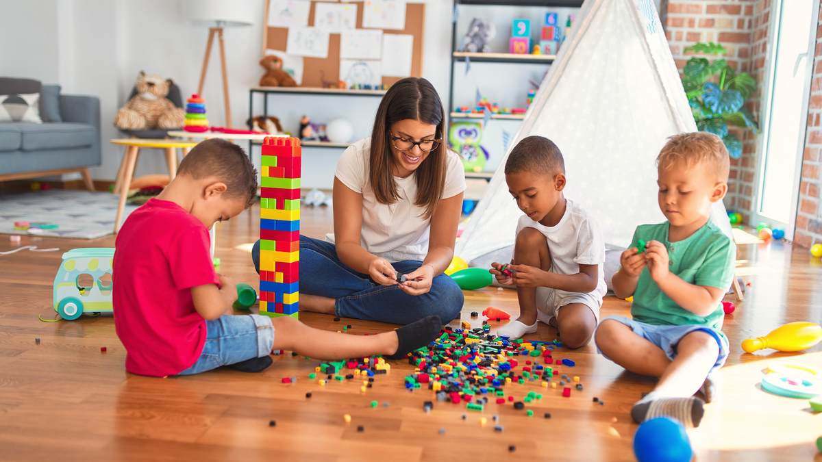 Professionnelle de la petite enfance assise au sol avec trois jeunes enfants, jouant avec des briques de construction