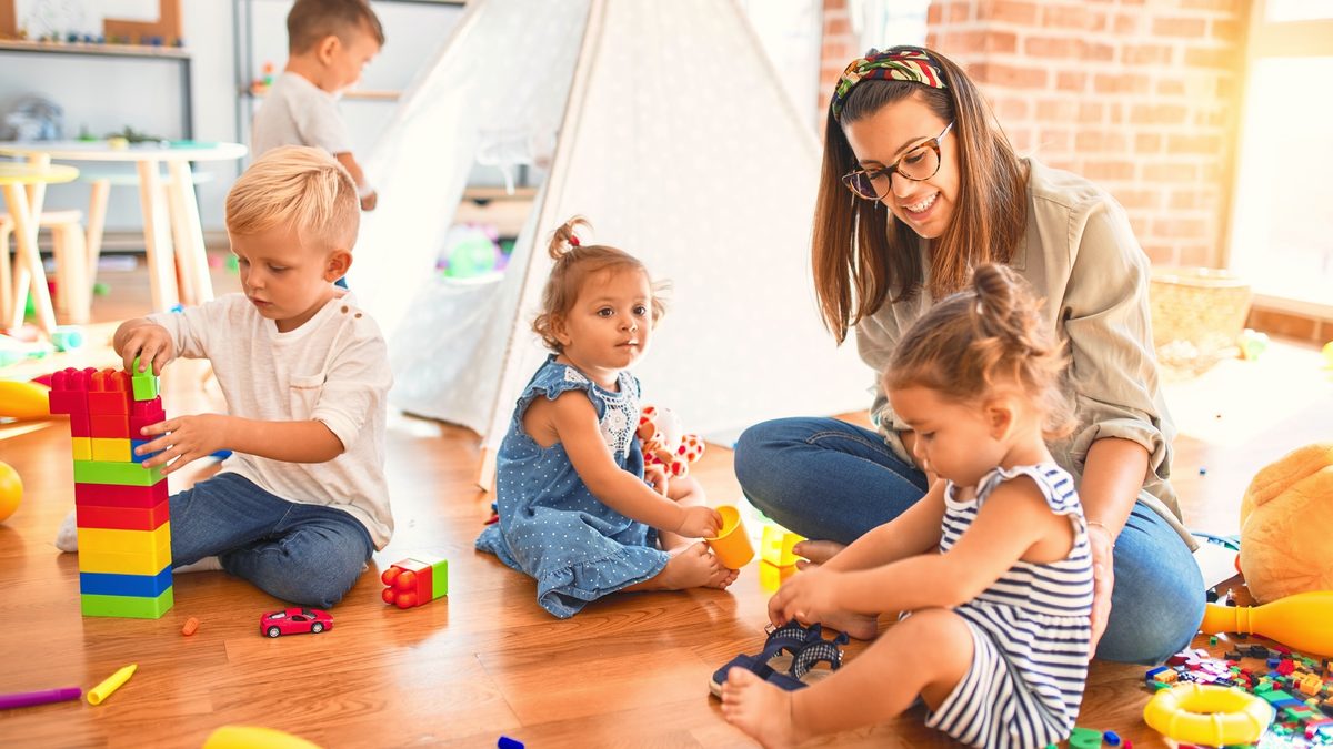 Une professionnelle assise par terre dans une salle de jeux de crèche, entourée de trois jeunes enfants qui jouent avec des cubes et des jouets