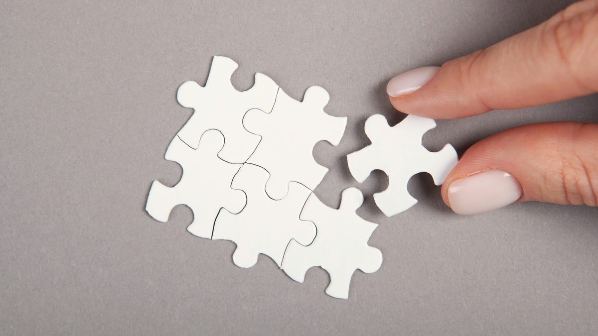 Une main pose la dernière pièce d'un puzzle blanc sur un fond gris, à côté de pièces déjà assemblées