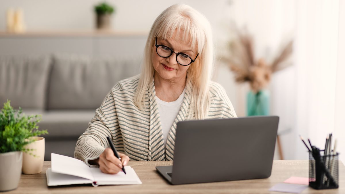 Femme assise à une table en bois, écrivant dans un carnet devant un ordinateur portable, dans un salon lumineux