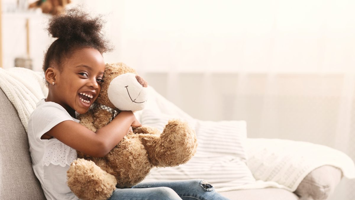 Une petite fille rit en serrant dans ses bras un grand ours en peluche sur un canapé clair